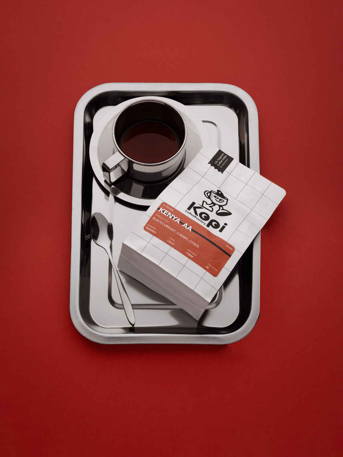 A top-down view of a coffee product presentation on a silver tray against a solid red background. The set includes a cup of black coffee on a saucer, a spoon, a box of Kopi coffee, and a stack of matching coffee bags.